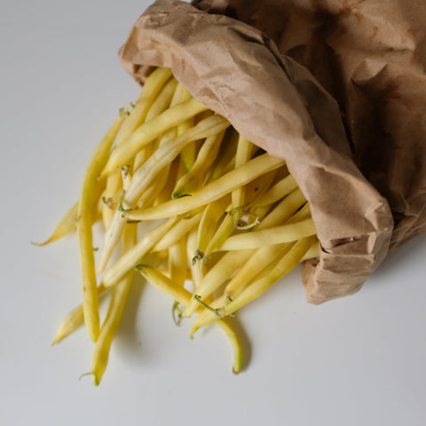 Yellow garden beans spilling out of a brown paper bag on a light gray background.